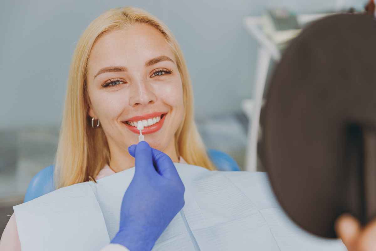 Young smiling calm happy woman choose veneers enamel color compare teeth shade with stomatologist look at result mirror sit at dentist office chair indoor modern cabinet. Enamel whitening treatment.