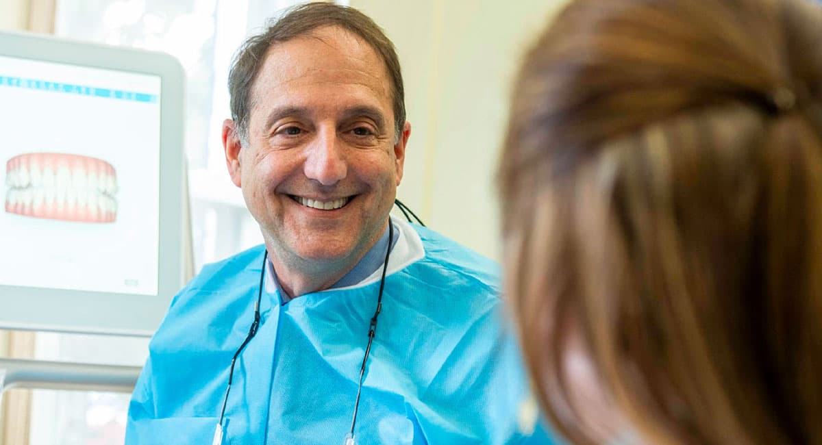 A smiling dentist in a blue protective gown and bib sits next to a dental patient, engaging in a friendly conversation. In the background, a dental monitor displays an image of teeth