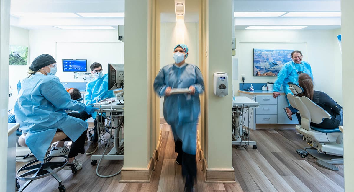 Dental professionals in protective gear attending to patients in our Olney dental office. One dentist is walking through a hallway carrying a tray, while others are engaged in procedures and consultations with patients.